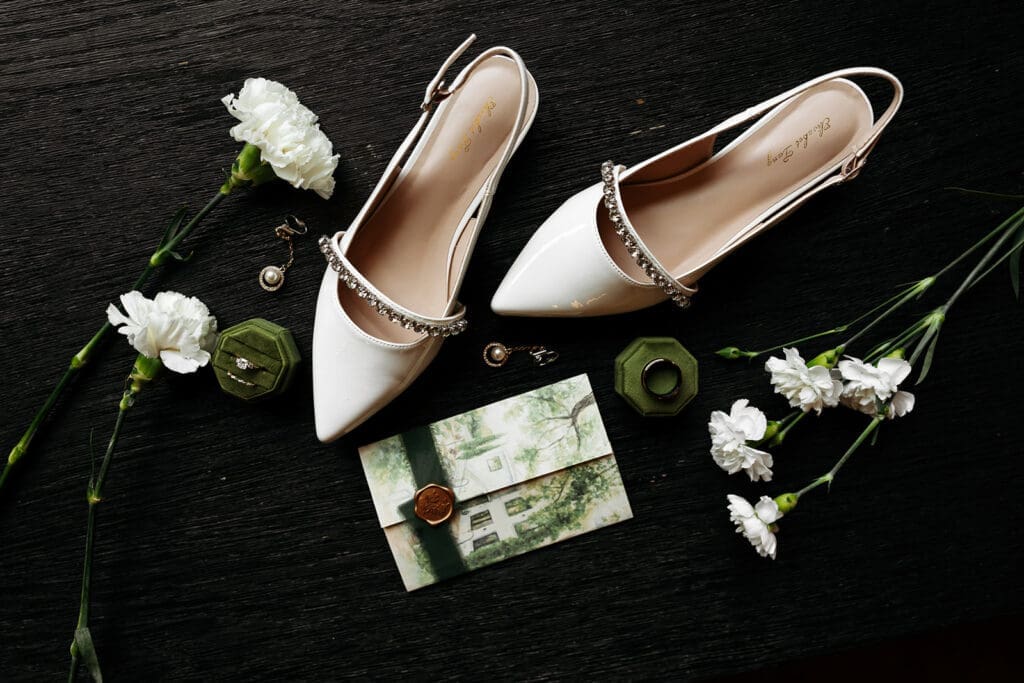 Bridal shoes, heirloom jewelry, wax seal, ring box, and white florals arranged in a refined wedding flat lay composition.