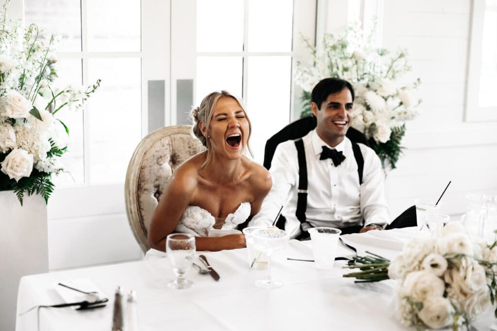 Bride and groom laughing during reception speeches surrounded by white florals and bright estate-inspired interiors.