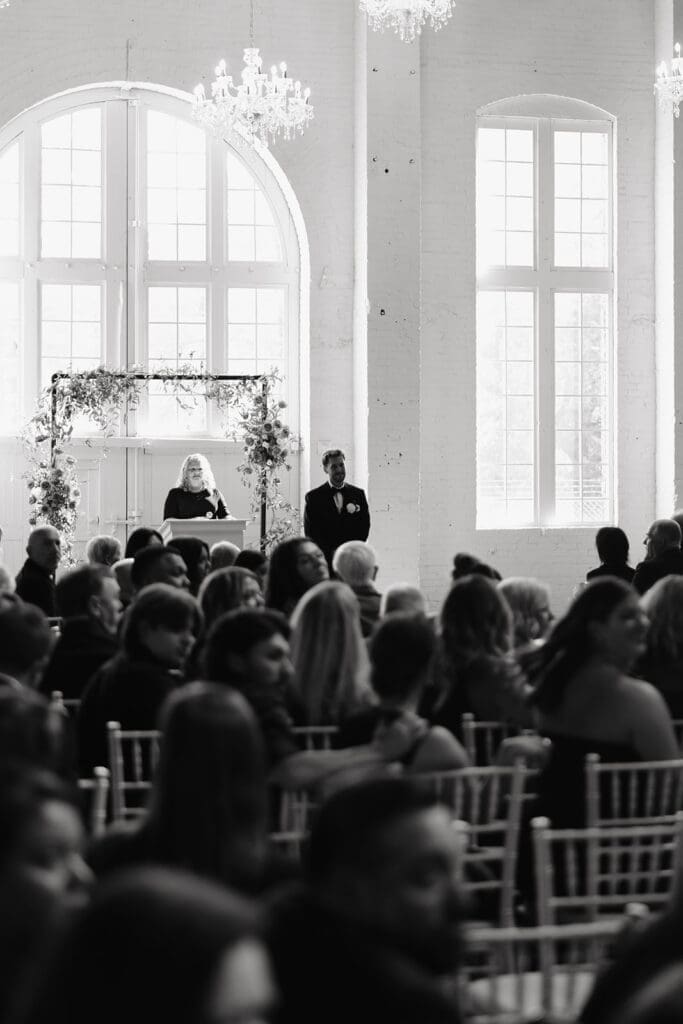 Ceremony view framed from the back of the room, showcasing the officiant, guests, and expansive architecture that adds depth to wedding photographs.