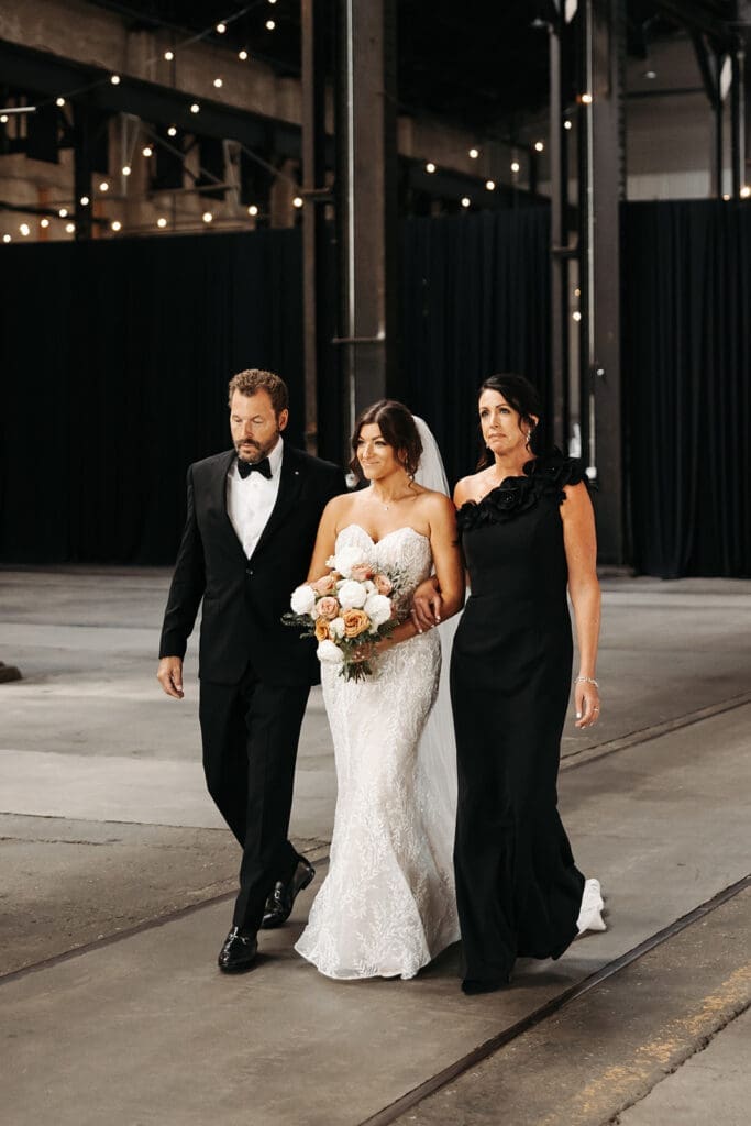 Bride walks down the aisle with her parents inside a historic industrial venue, demonstrating how longer aisles create more opportunities within wedding photography and videography packages.