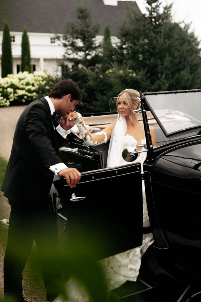 Groom helping his bride step out of a vintage getaway car after portraits at a vineyard wedding venue.
