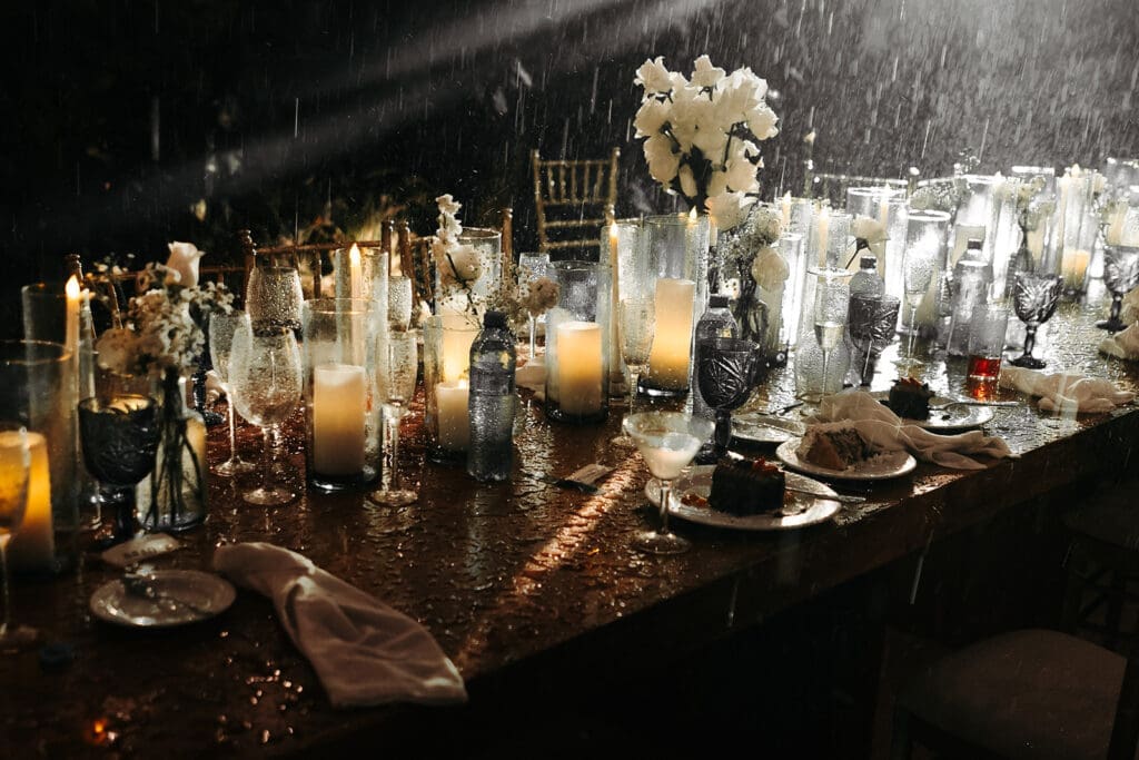 Rain-soaked reception table glowing with candlelight, layered glassware, and romantic evening wedding styling.