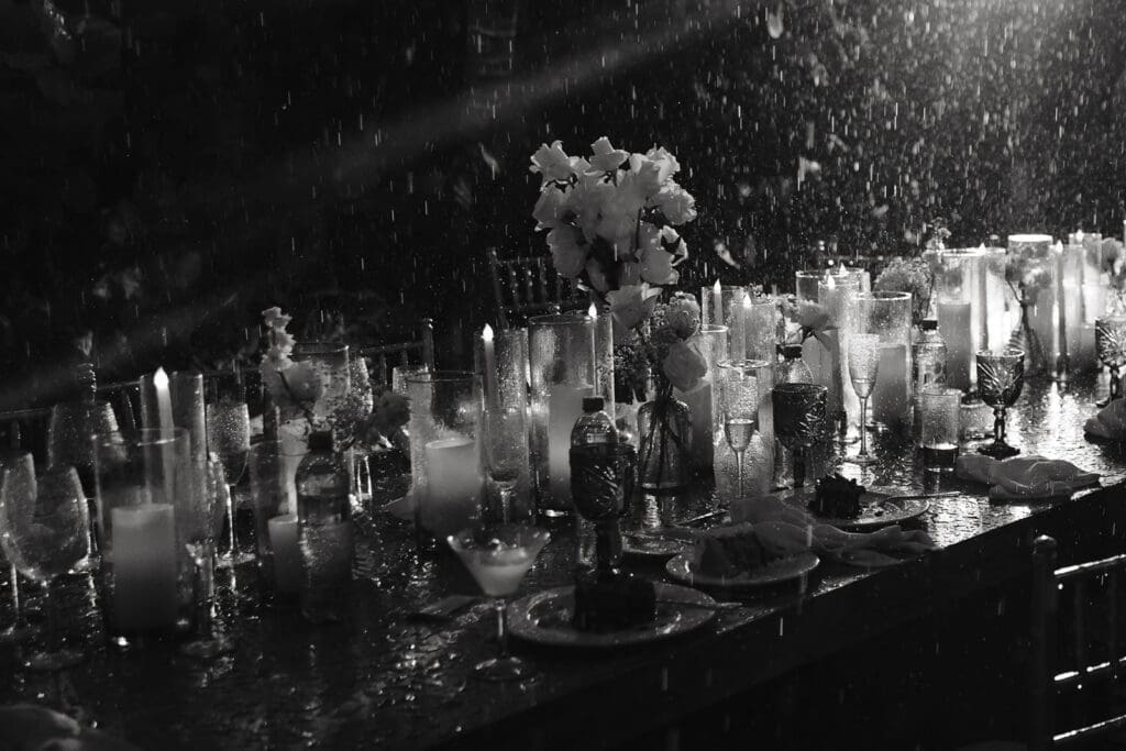 Rain-covered reception table glowing with candlelight and glassware during an atmospheric evening wedding celebration.