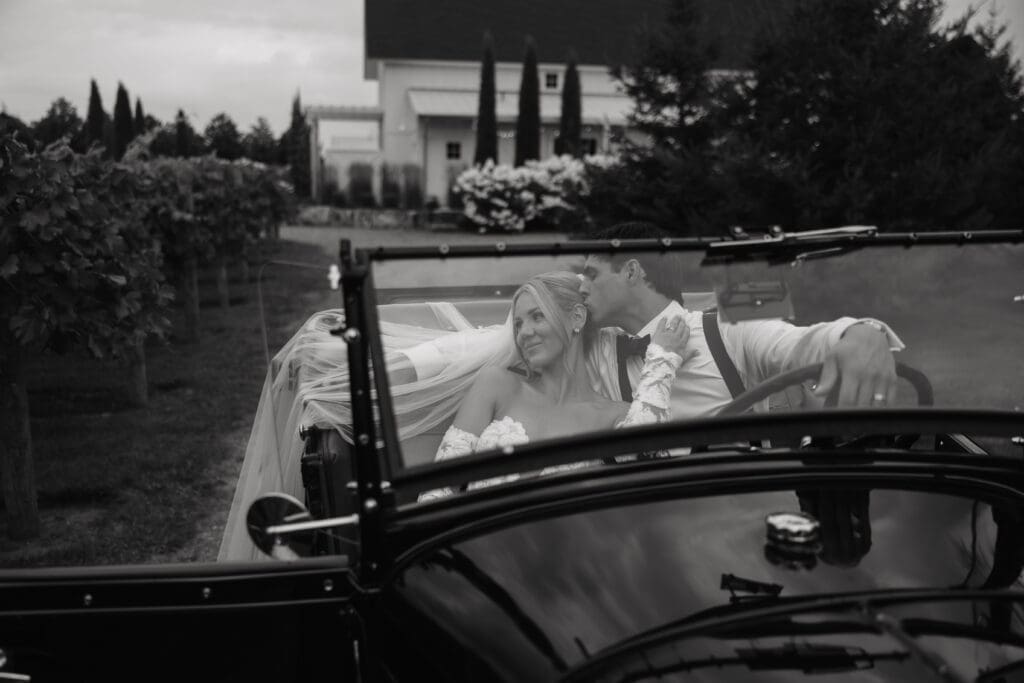 Bride and groom cuddled together in a vintage getaway car with the vineyard and white barn in the background.