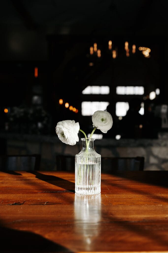 Single white ranunculus displayed in a ribbed glass vase on a sunlit reception table.