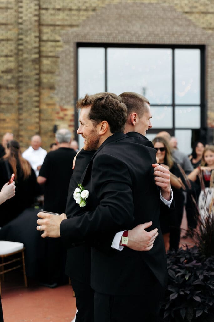 Groom hugs a friend during cocktail hour while guests celebrate after first look photos.