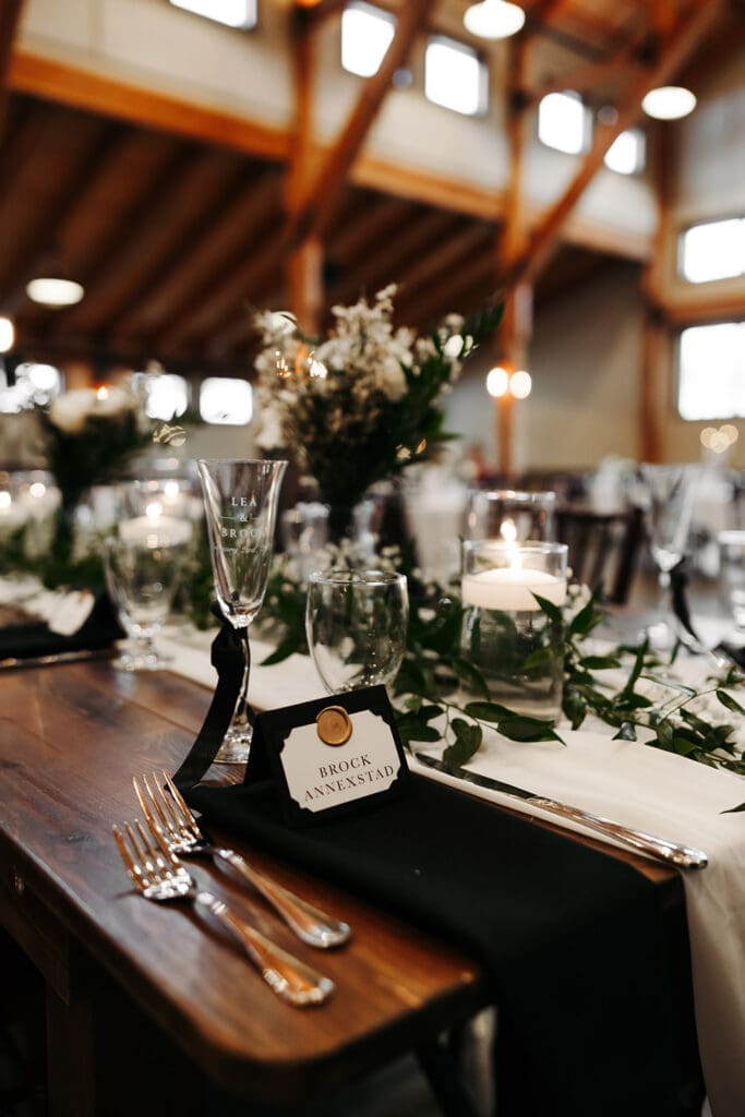 Reception place setting featuring black linens, candlelight, greenery, and a personalized guest name tag at a farmhouse-style table.