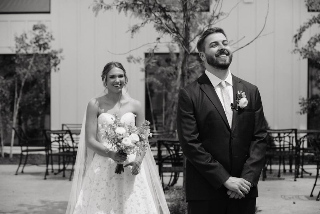 Groom laughs through happy tears during his wedding first look while the bride watches from behind.