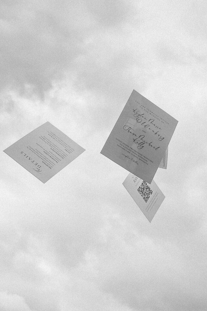 Creative wedding detail photo of an invitation suite photographed midair against a cloudy sky, showcasing modern stationery design.